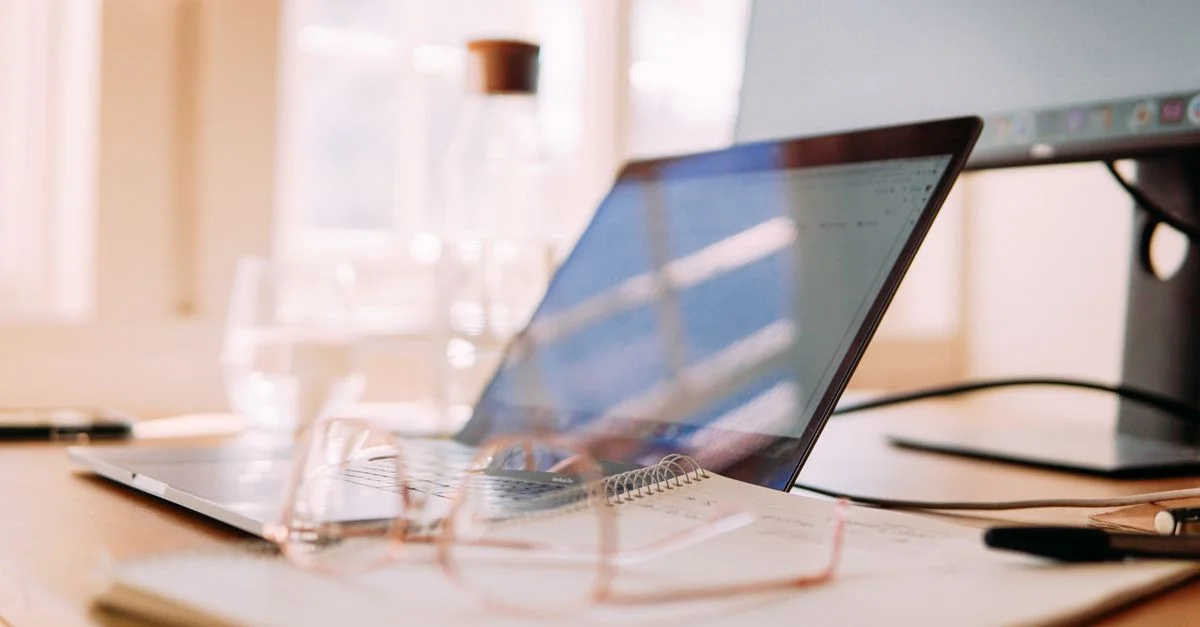 Un espace de travail à domicile bien éclairé avec un ordinateur portable, un écran et des lunettes, idéal pour le télétravail.